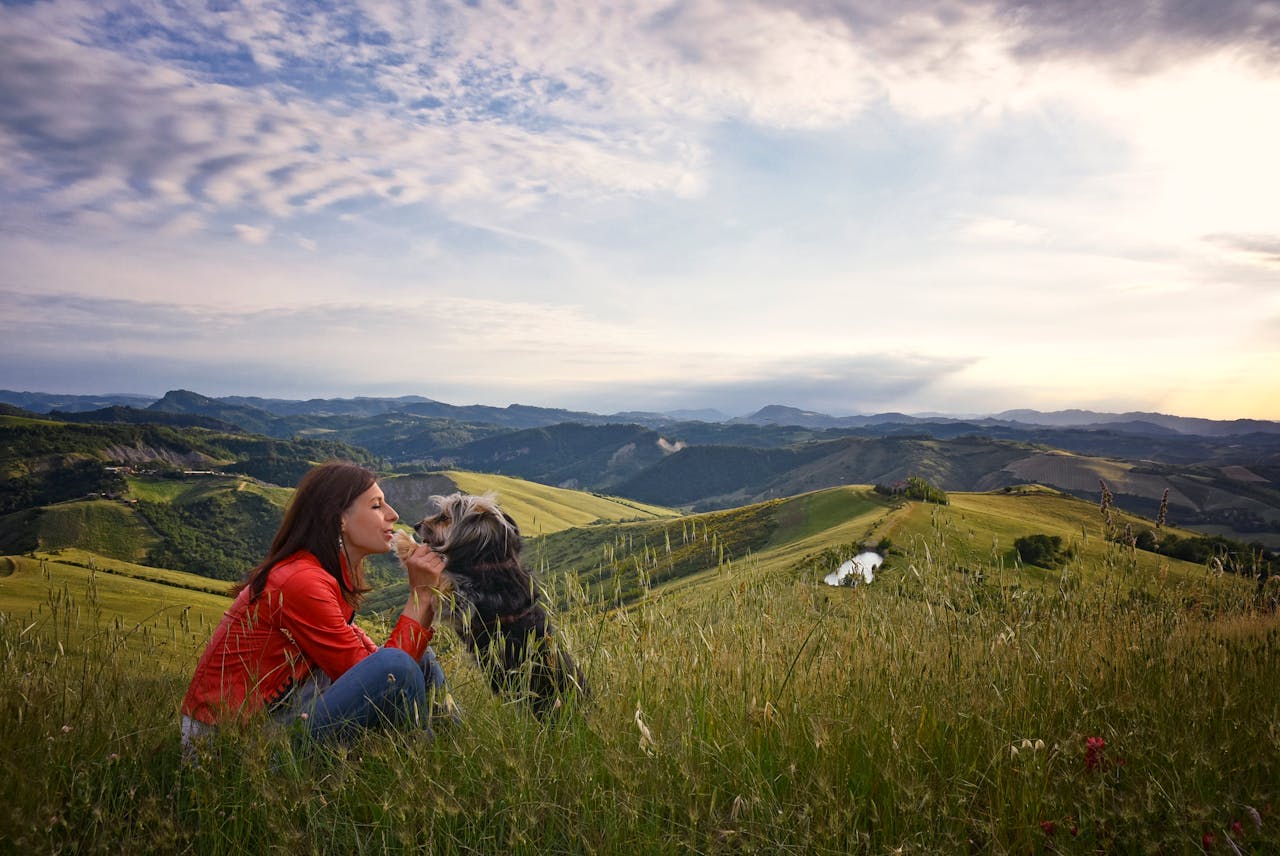 A woman sits with her dog on a grassy hillside, embracing natures beauty under a cloudy sky.