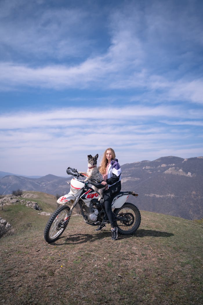 A young woman with a dog sits on a motorcycle atop a scenic hill.